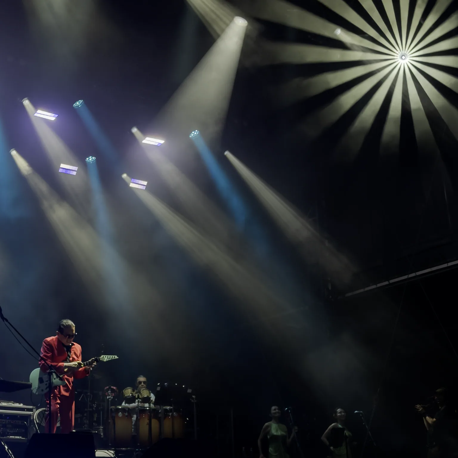 Masayoshi Takanaka playing guitar at the edge of a dark stage beneath sweeping beams and a radial white light.
